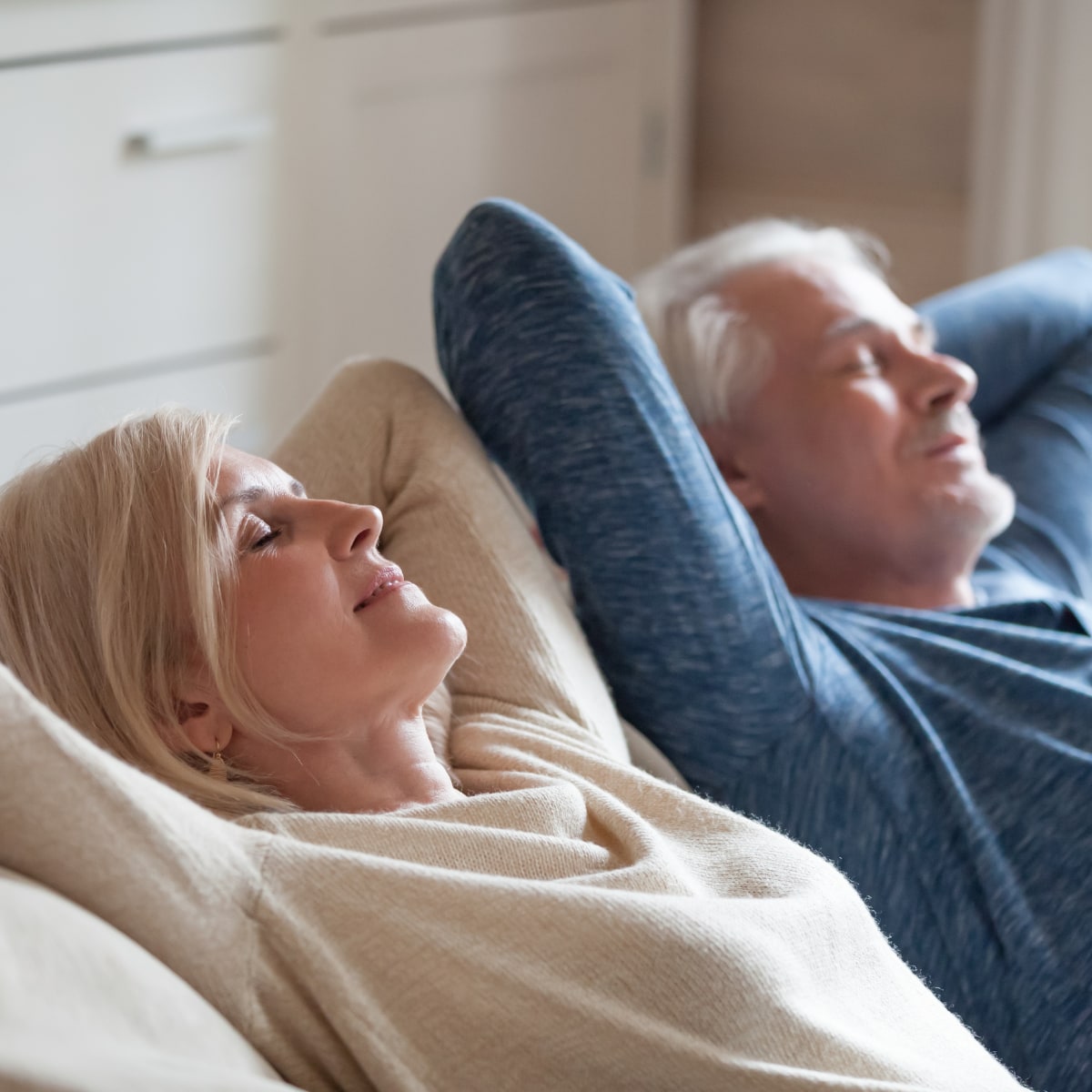 older mand and woman lounging back on a couch relaxing with their arms back cradling their heads