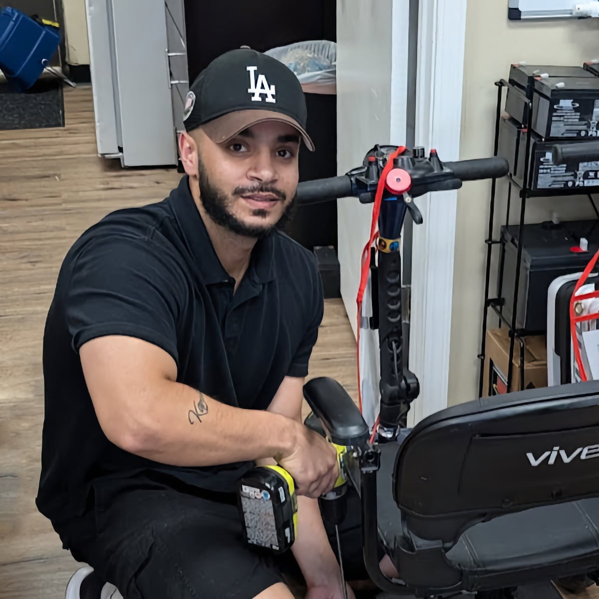 Taha; Midwest Mobility Field Service Technician. Young man with baseball cap kneeling near a scooter