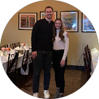 Benjamin Sonnenburg and a woman stand together, smiling broadly in a restaurant setting with formal dining tables behind them.