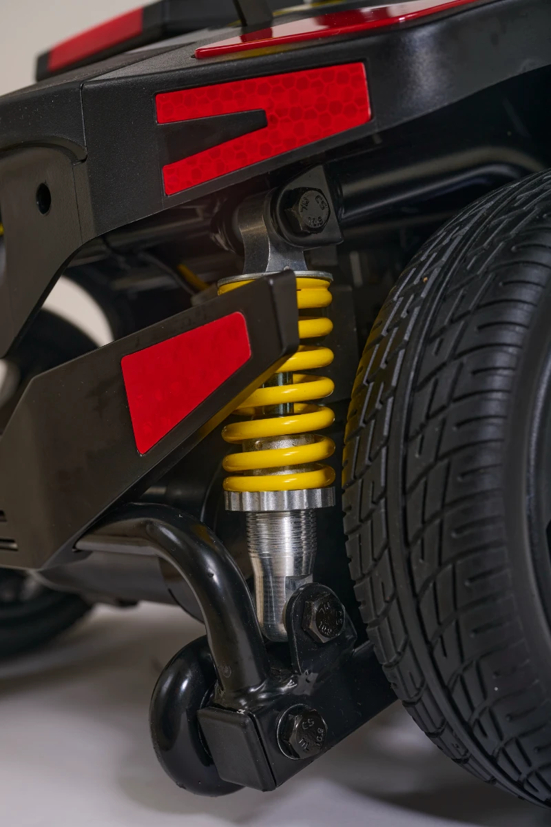 A close-up of a mobility scooter’s suspension system, showing a bright yellow coil spring shock absorber next to a black tire. Red reflectors accent the black chassis.