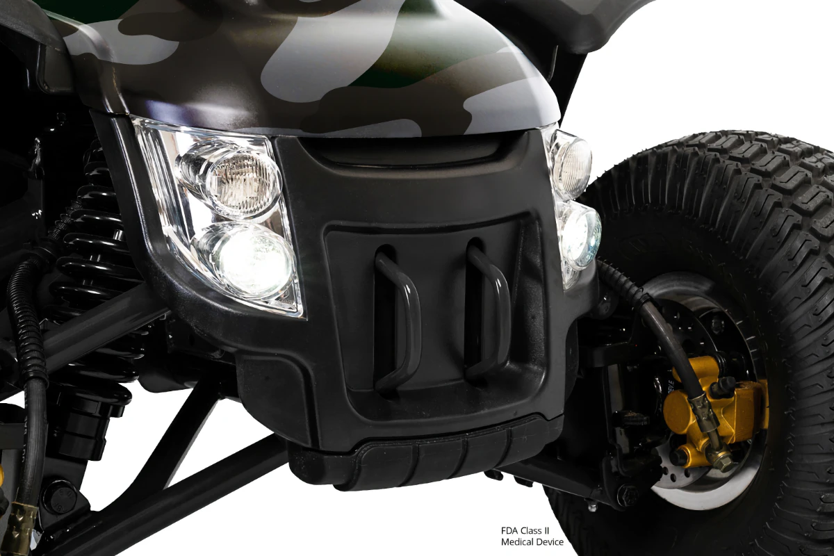 Close-up of a camouflage mobility scooter's front, featuring bright dual headlights, heavy-duty suspension, and a large, knobby off-road tire.