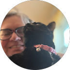 Cyan Kuehl, wearing glasses and smiling, holds a black cat with a pink collar looking up affectionately at her.
