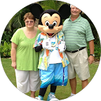 A happy couple stands posing with Mickey Mouse, dressed in a Hawaiian shirt and shorts, outside against a backdrop of tropical greenery.